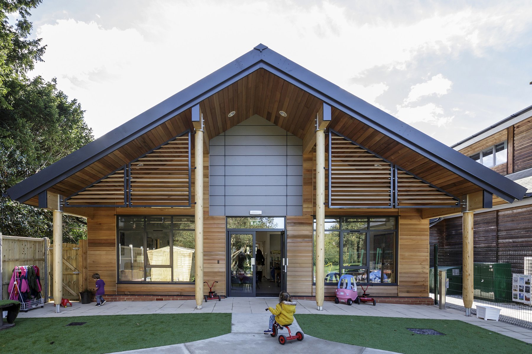 The finished nursery from the play area: a broad cedar gable on round timber columns, with louvred screens either side of the entrance