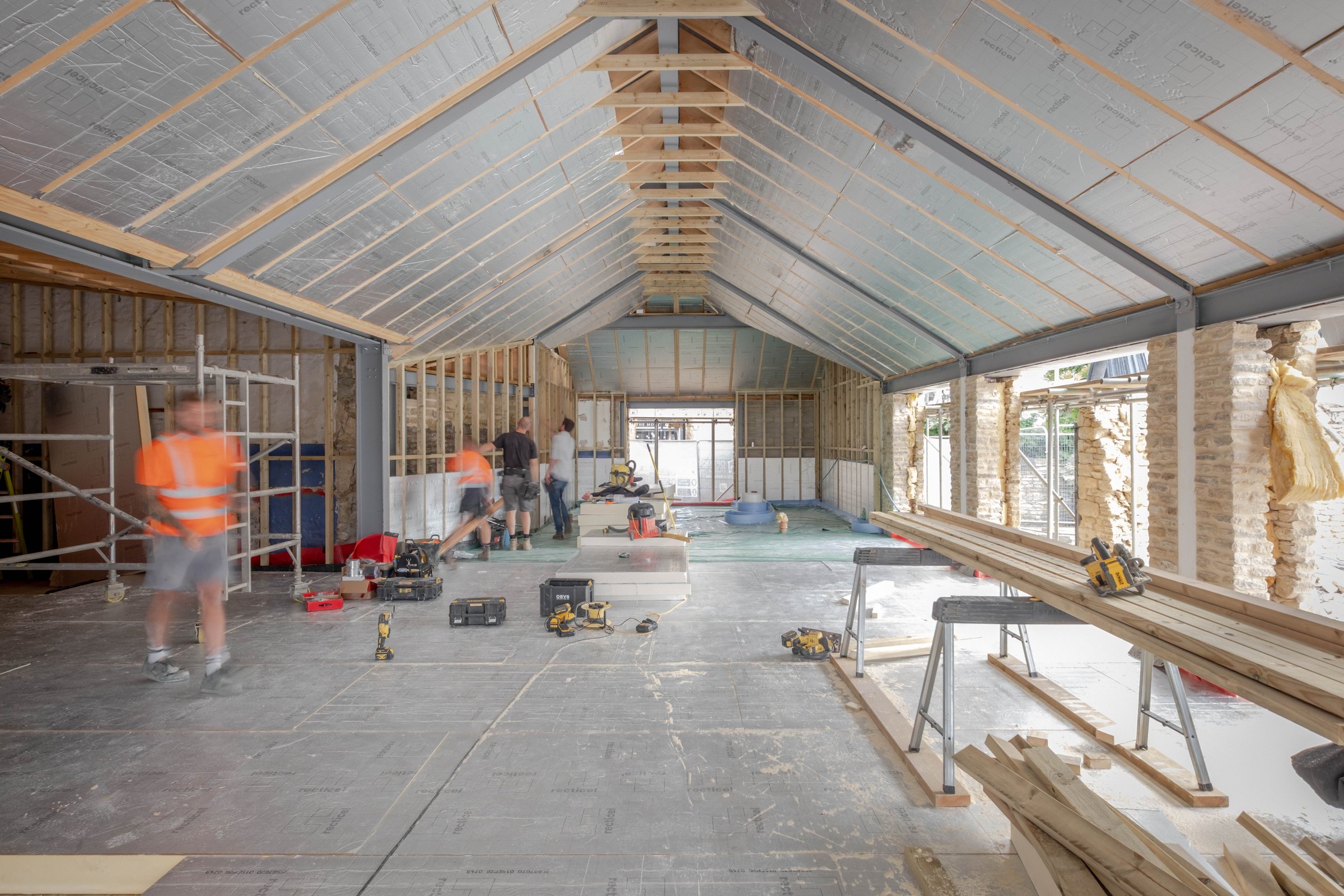 The new workshop at Market Square during construction: timber roof structure and insulation above, old stone walls to one side, trades at work